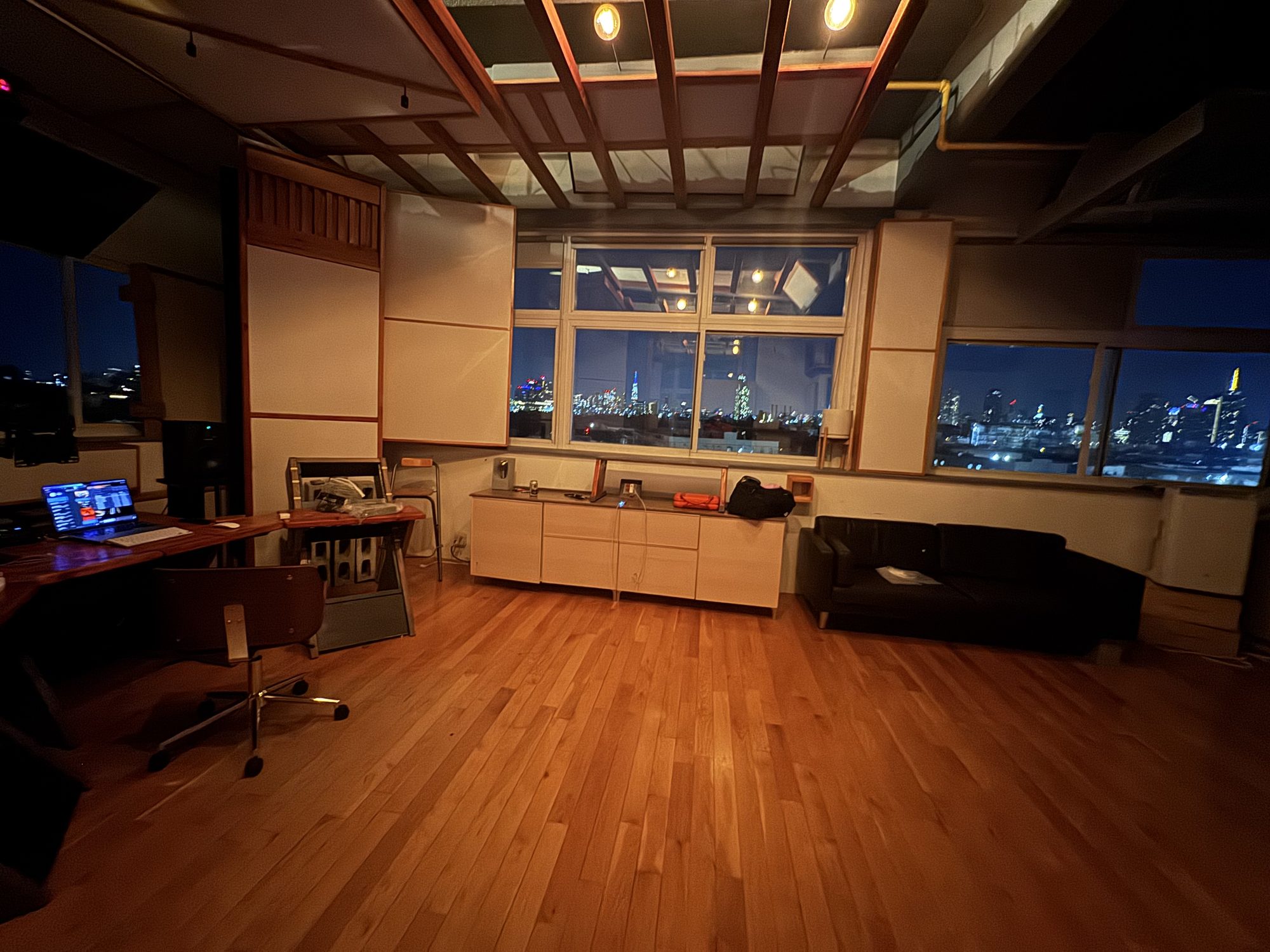 The live room at night: timber ceiling grid, panels between the beams, a black sofa, and the Manhattan skyline through the windows.